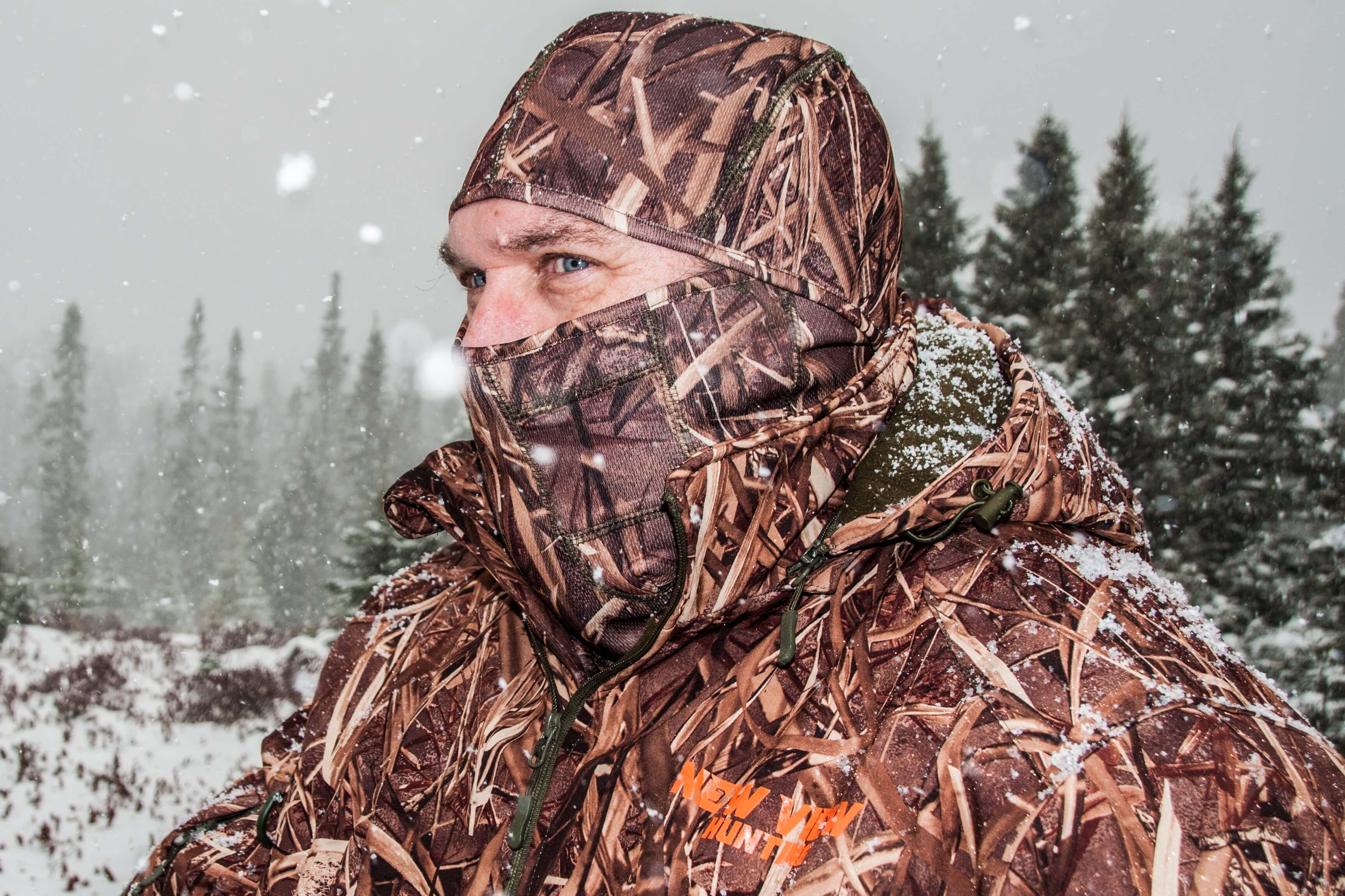 Hunter wearing camouflage face covering and cold-weather hunting accessories in snowy woodland